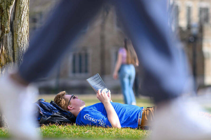 A student lies in the grass reading on a warm day as another student walks by in the foreground.