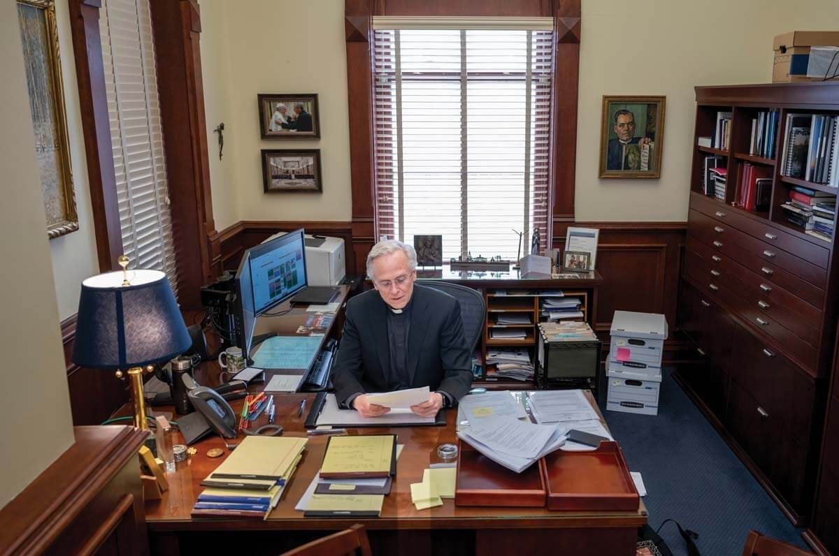University President Rev. John I. Jenkins reviews a paper at this desk, which is covered with several other notebooks and documents.