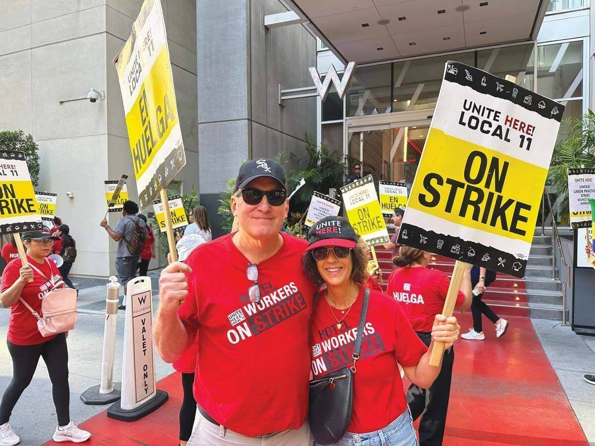 Kurt Petersen and his wife, Anne DeWald Petersen, wear red shirts and hold yellow picket signs at a demonstration for hotel workers' rights.