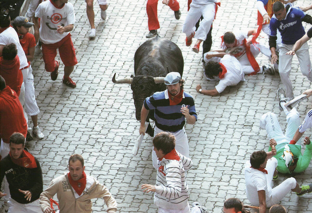 Bill Hillman, in a black and blue striped shirt with a red bandana around his neck, runs just steps ahead of a bull in the middle of a crowd in Pamplona, Spain.