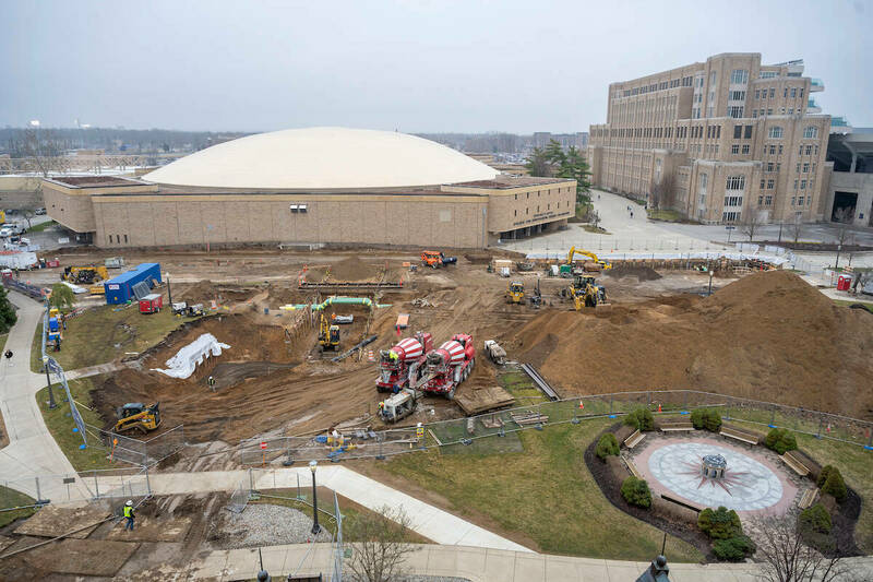 A hole in the ground, next to a dirt mound with construction equipment in place near the white, north dome of the Joyce Center.