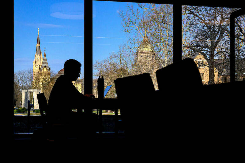 A person in silhouette sits in front of a laptop at a library table against a bright blue sky and sunshine through the windows with the Golden Dome and basilica in the background.