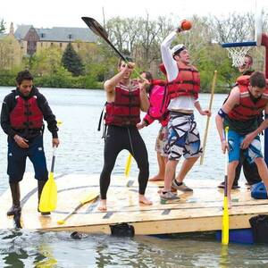 Students competing in the Fisher Regatta on St. Mary's Lake play Nerf basketball on their boat, with one going up for a dunk.