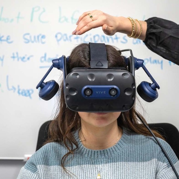 A student sits in front of a whiteboard with writing on it, wearing a virtual reality headset with a hand hovering above her head, ready to help.