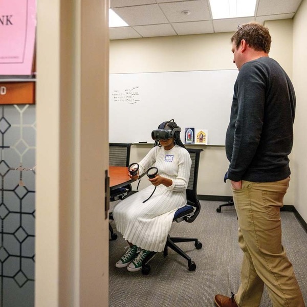 A professor walks through the open door of a classroom with a student seated inside wearing a virtual reality headset.