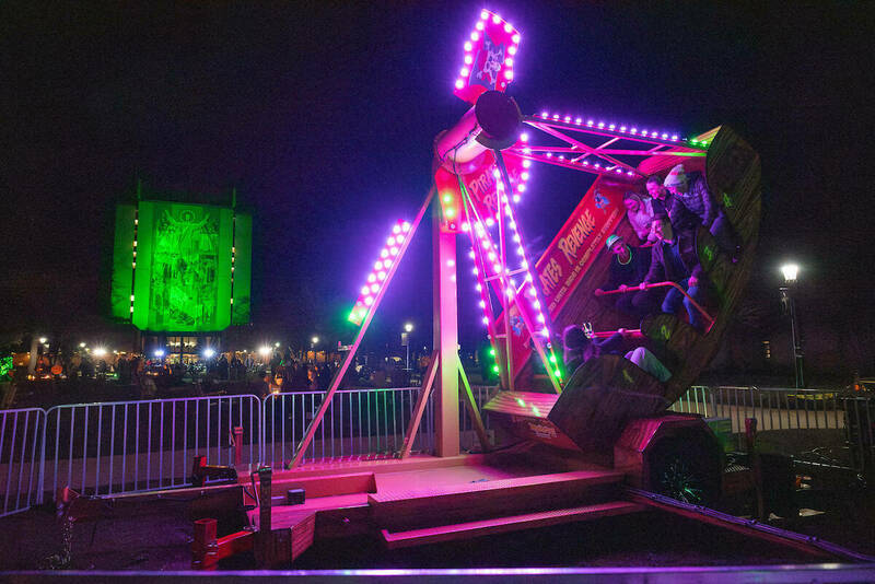With the Touchdown Jesus mural illuminated in green in the background, a carnival ride lit up in pick and purple swings on the Library Lawn.