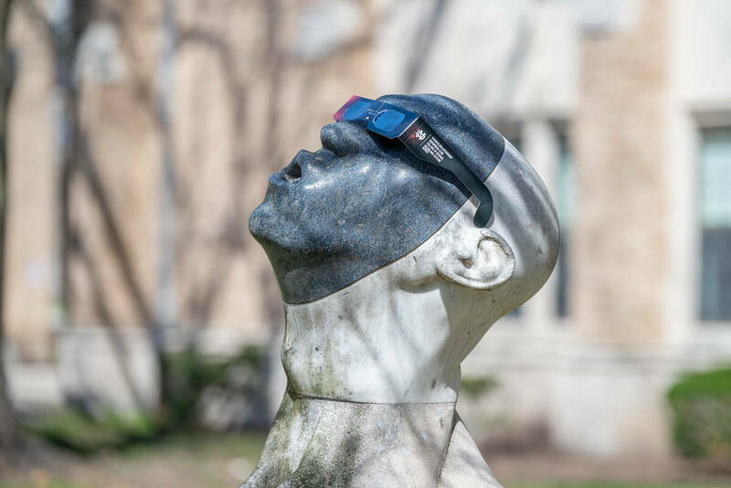 A scuplture of a head with its neck craned upward wears eclipse glasses, appearing to be taking in the phenomenon.