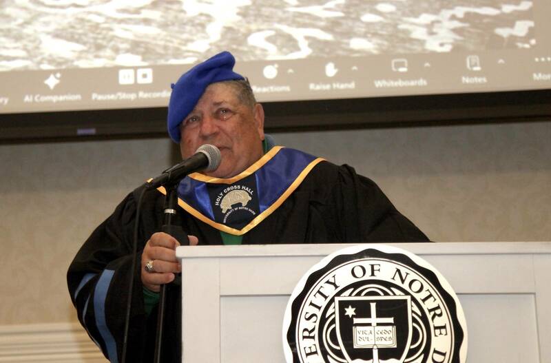 Drew Danik, dressed in academic regalia, speaks into a microphone after receiving an honorary doctory of reunionology degree for his work to keep residents of Holy Cross Hall in touch over the years.