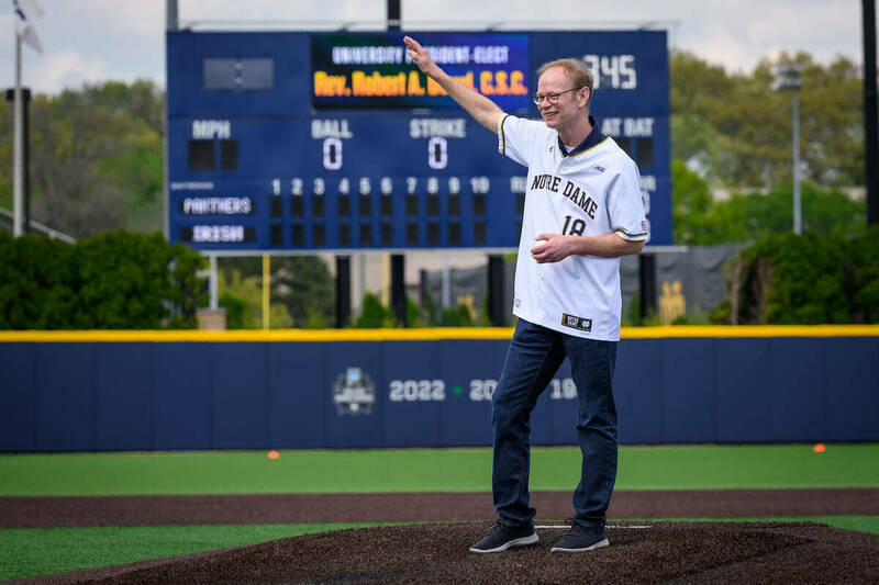 Father Bob Dowd, wearing a notre Dame basball jersey and blue jeans, waves to the crowd while standing on the mound, preparing to throw out the first pitch at a Notre Dame baseball game.