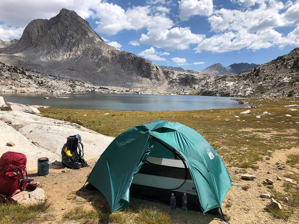 A tent in the mountains under blue skies with a few clouds and near a lake along the John Muir Trail.