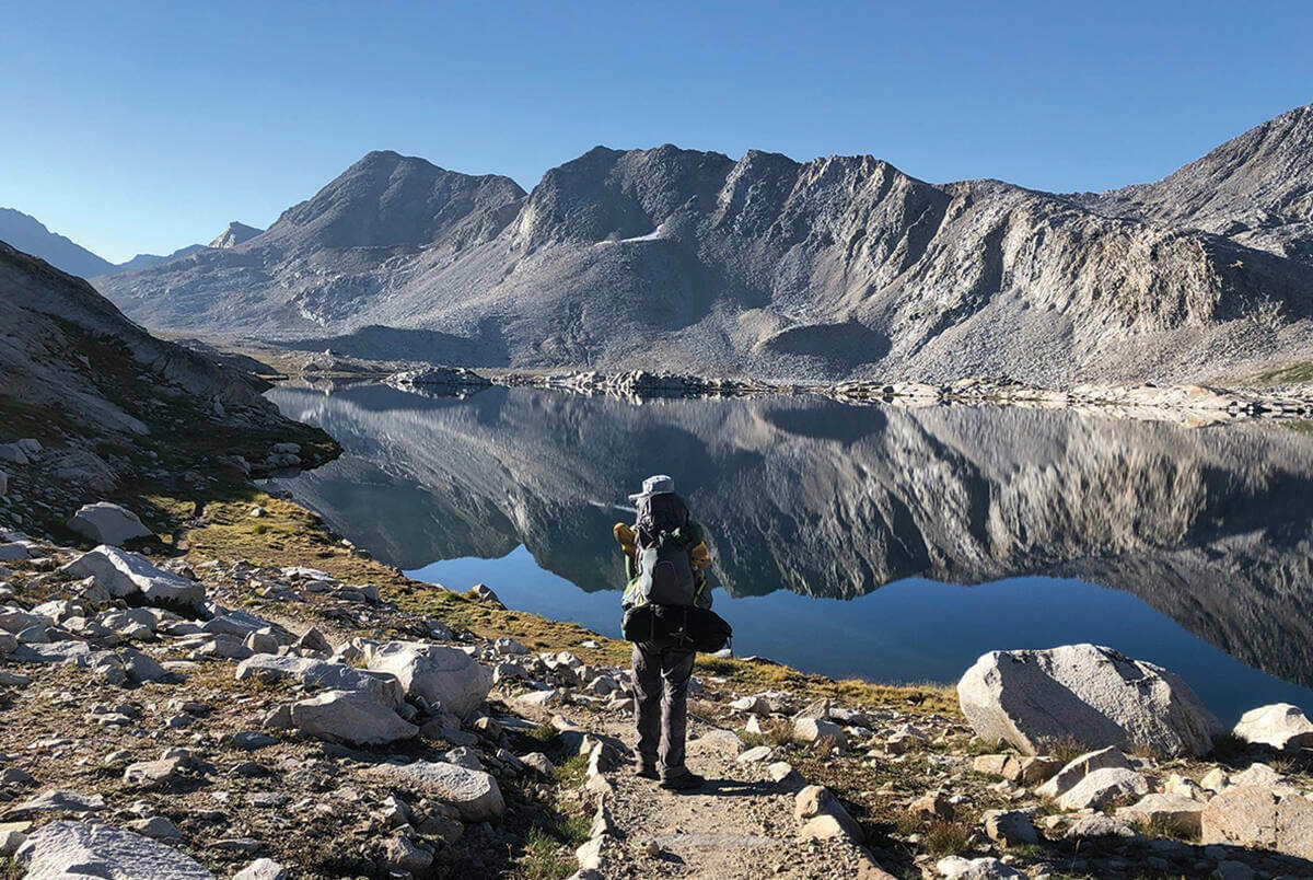 A person hikes toward a lake with mountain peaks in the distance along the John Muir Trail.