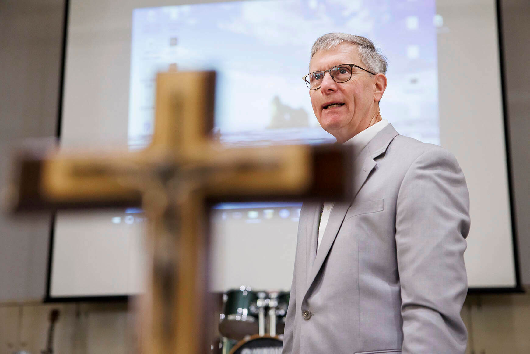 Deacon John Cord speaks with an image on a screen behind him and a cross in the foreground