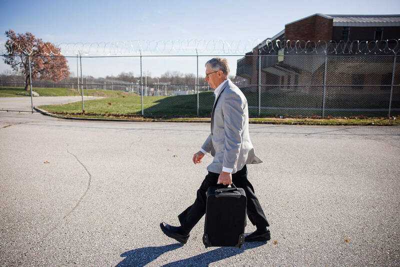 Deacon John Cord walks in front with a barbed wire fence and a prison building behind him.
