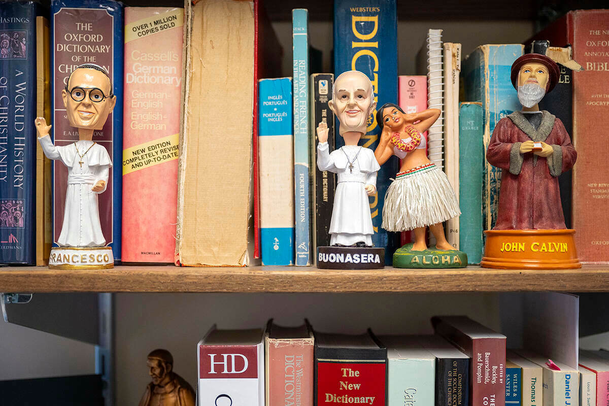 Books and bobblehead dolls of religious figures and a hula dancer on a shelf in William Cavanaugh's office