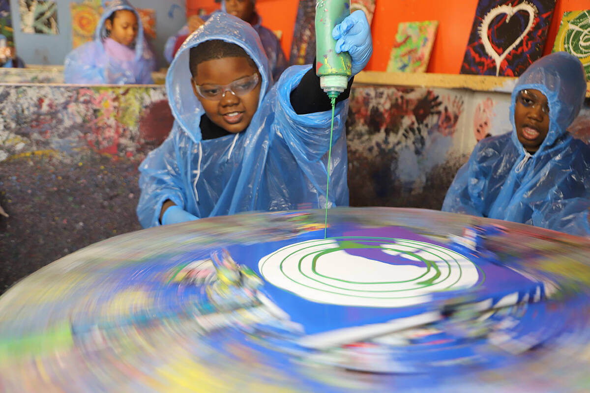 A young student wearing a poncho smiles as he pours paint on a spinning wheel during a Horizons for Youth outing