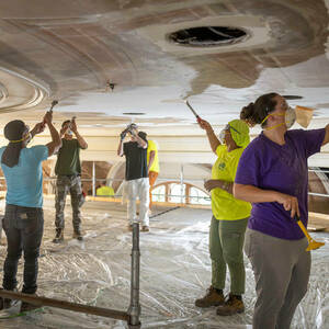 Six people are shown scraping paint from a ceiling.