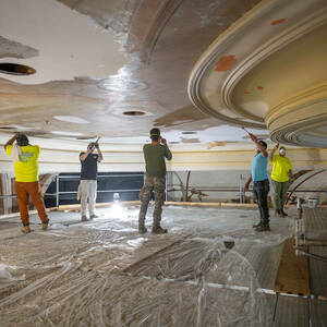 Six workers are shown scraping paint from a ceiling.