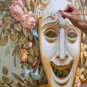 An artist's hand holding a paintbrush puts detailed touches on a six-foot canvas painting of a mask representing comedy to be hung in Washington Hall.