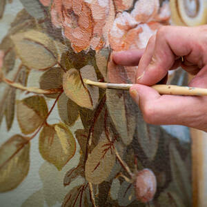 An artist's hand holding a paintbrush dabs bits of paint to a leaf design on a wall mural.