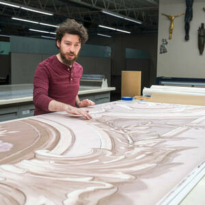 A young man with a beard and curly brown hair stands in front of a table covered with ornate etchings.