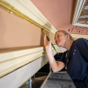 A young woman uses a paint brush to smooth shiny gold leaf along the wooden surface of a wall.