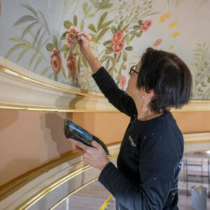 A woman holding a paint brush in her right hand paints images of flowers and grasses on a curved ceiling.