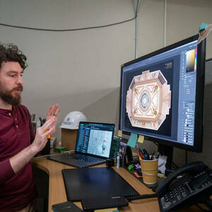 A young man with a beard and curly dark hair gestures with his hands before a computer screen.