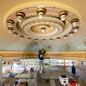 A woman stands on a scaffolding painting an image of flowers and grasses on the curve of a domed ceiling.