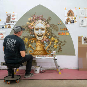 A man facing a wall paints an ornate mural showing the smiling face symbolizing comedy theater.