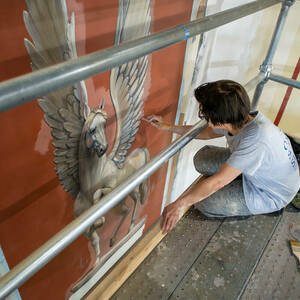 A woman sits on a scaffolding and paints details on a wall mural of a winged horse.