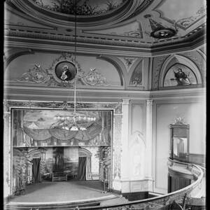 A black and white image showing a stage and walls and part of a ceiling of an ornately decorated Victorian era theater, with a portrait of George Washington above the stage.