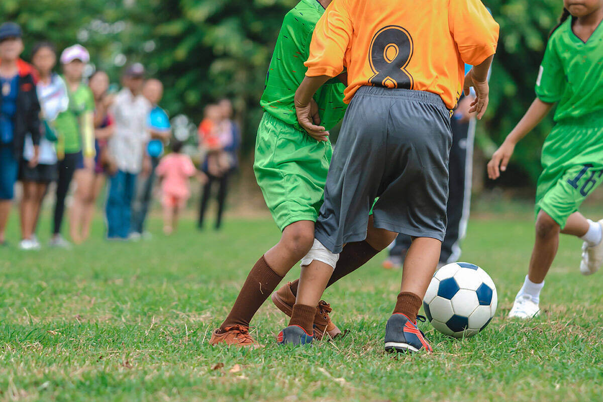 Young kids in orange and green jerseys chase a soccer ball while parents, blurred in the background, watch the game from the sidelines