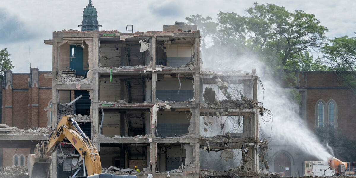 The shell of a mostly demolished Fisher Hall with dirt mounds and heavy equipment in front of the building with its side torn away to see through what once were rooms and hallways.