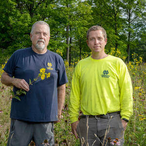 Two men, one hold a stem bearing yellow blossoms, stand in a field of native wildflowers.