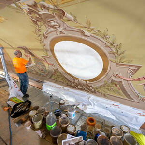 A man stands on a scaffolding painting an elaborate and colorful mural above an auditorium stage.