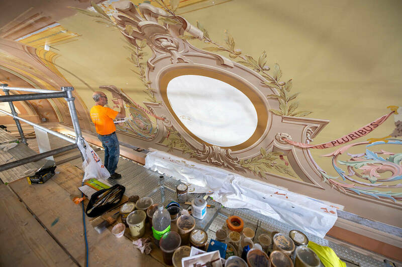 A man stands on a scaffolding painting an elaborate and colorful mural above an auditorium stage.