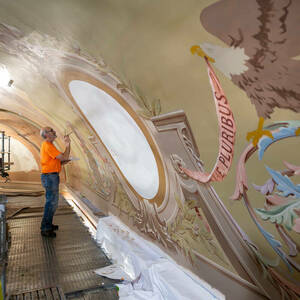 A man standing on scaffolding with a brush in his right hand paints an elaborate pattern on a curved wall.