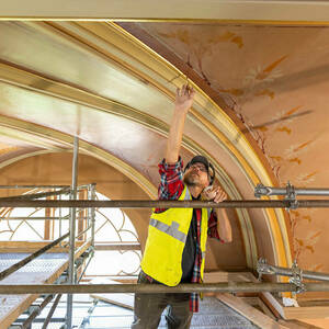 A man stretches his right arm high to a curved ceiling to apply paint with a narrow brush.