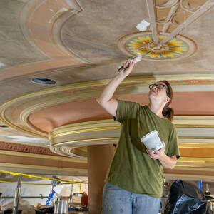 A woman with right arm raised patches cracks on a ceiling covered with colorful decor.