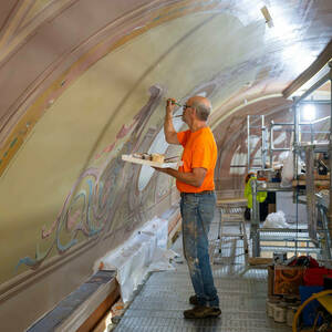 A man standing on a scaffolding applies small bits of paint to a curved wall from a narrow paintbrush he is holding in his right hand.