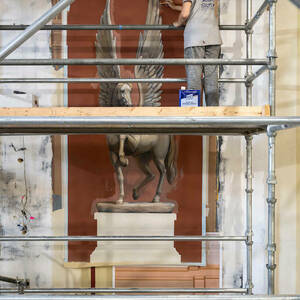 A woman stands on a scaffold painting an image of a large winged horse on a wall.