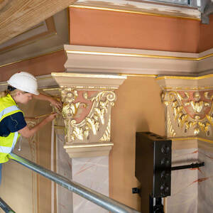 A woman wearing a construction hat uses both hands to apply gold material to the top of a pillar inside a building.