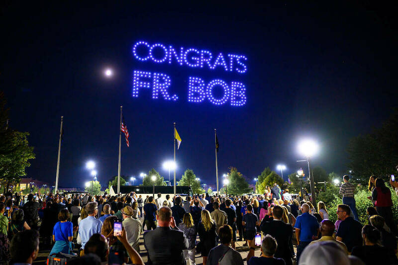 Illumniated drones spell out "Congrats Fr. Bob" in the night sky