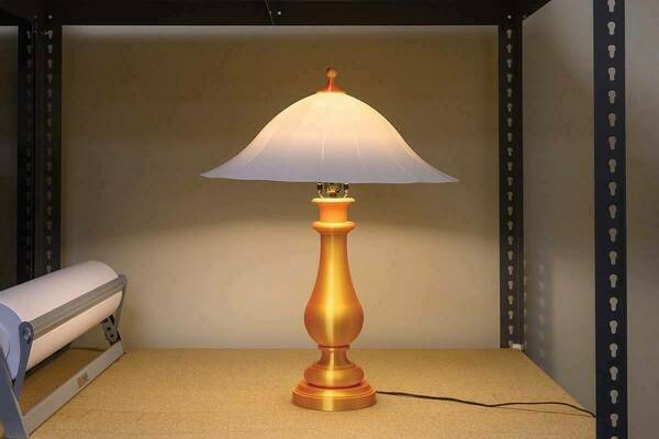 Gold-colored table lamp with a white flared shade, illuminated and sitting on a cork tabletop.  A paper roll dispenser is partially visible in the background.