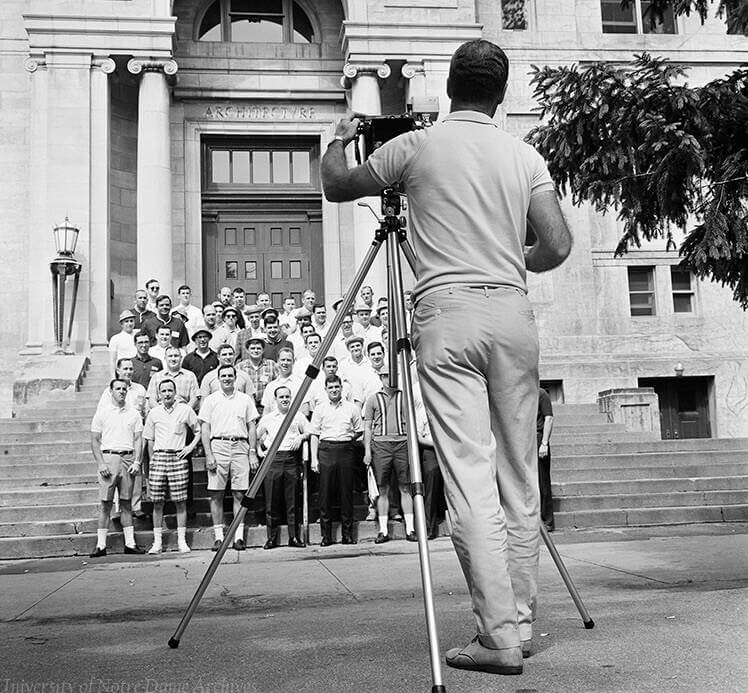 A man with a camera on a tripod photographs a large group of men on the steps of the Architecture Building at the University of Notre Dame.