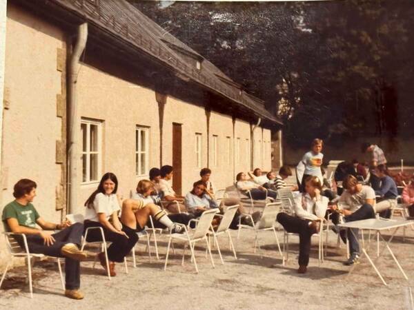 Students relax in white lawn chairs outside a tan building with a dark brown shingled roof.  Some students wear shorts and knee socks.  One student wears a green t-shirt.  Another wears a horizontally striped shirt.