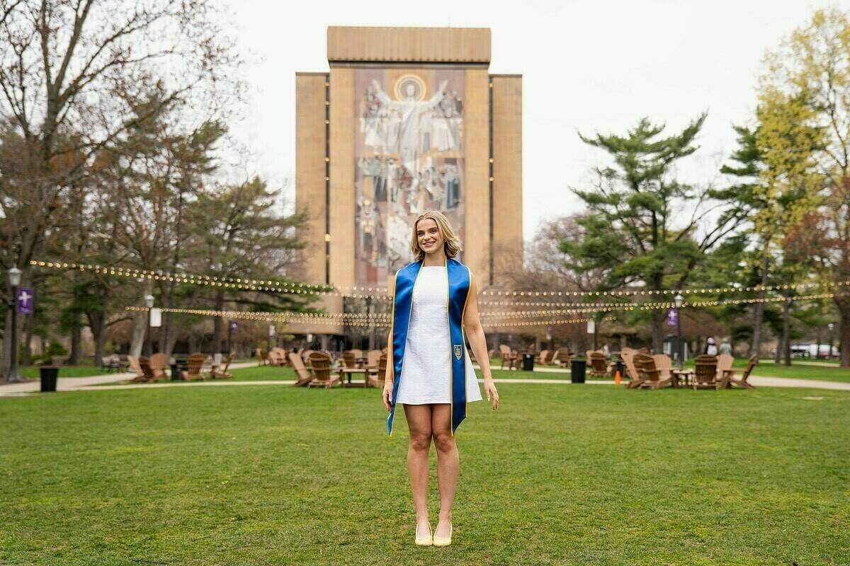 A blond graduate wearing a white dress and a blue and gold stole smiles on the lawn in front of Hesburgh Library at the University of Notre Dame.