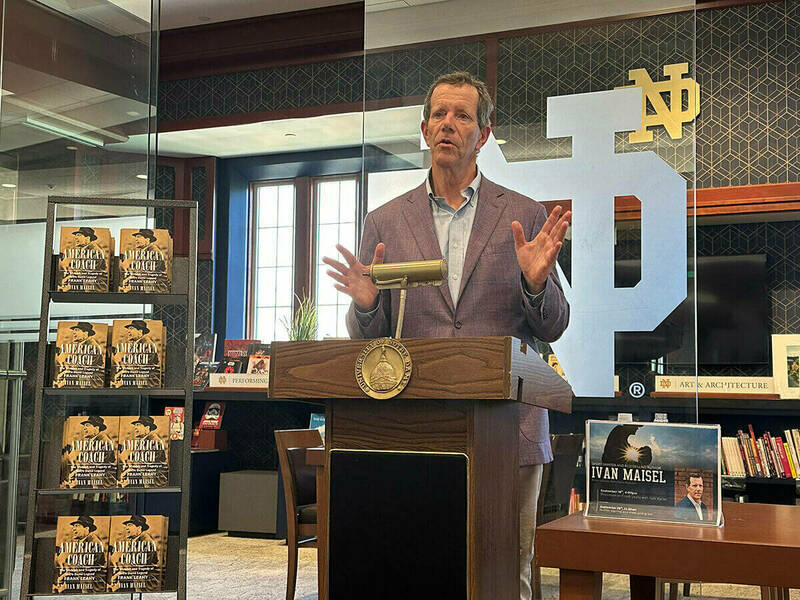 A man speaks at a podium, gesturing with open palms, next to a display of books.