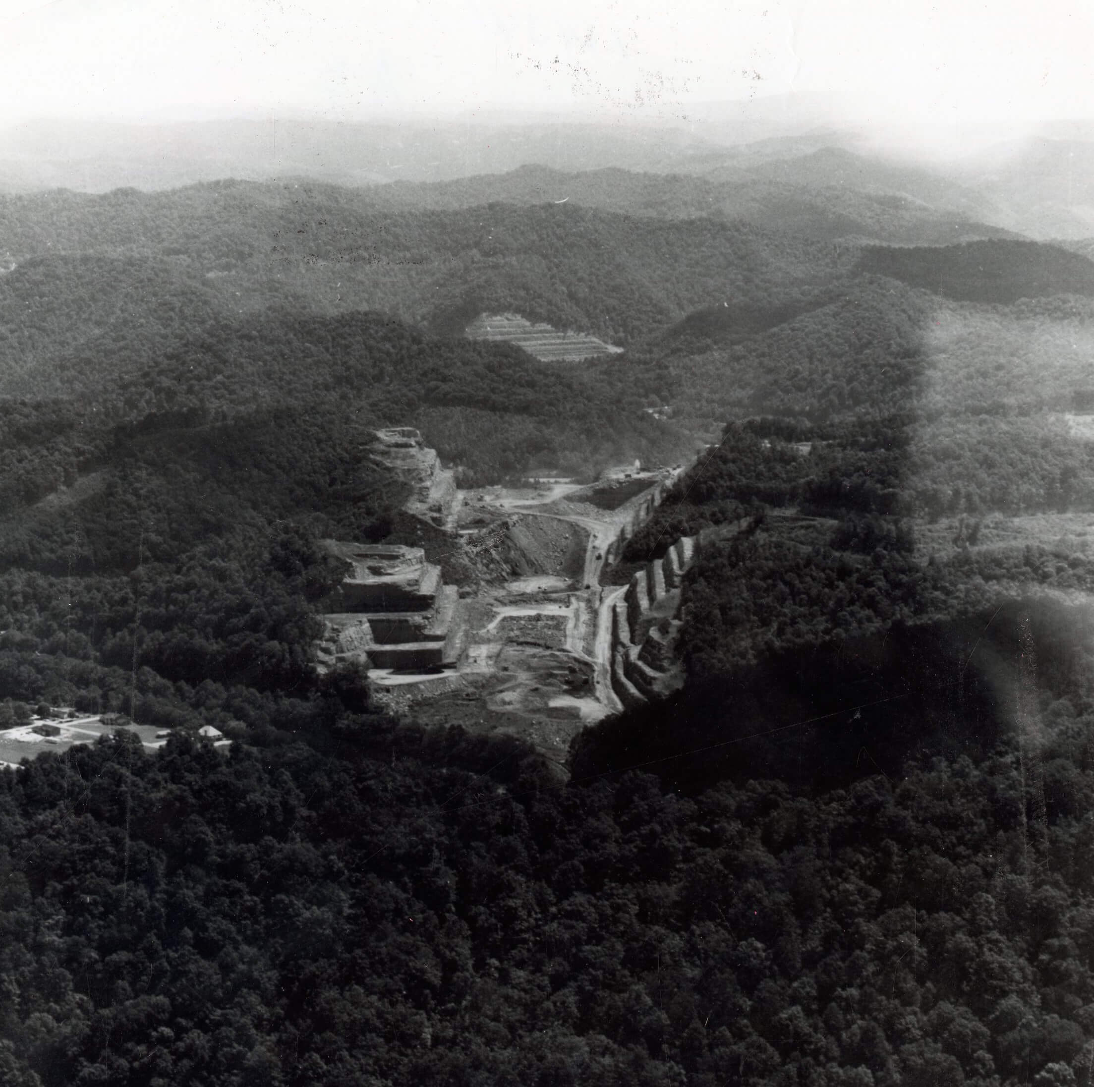 Black and white aerial view of a vast terraced excavation carved into a dense, forested mountain range. Unpaved roads wind through the barren ground, with heavy machinery visible. A small cluster of buildings lies at the lower left, surrounded by trees and distant hazy mountains.