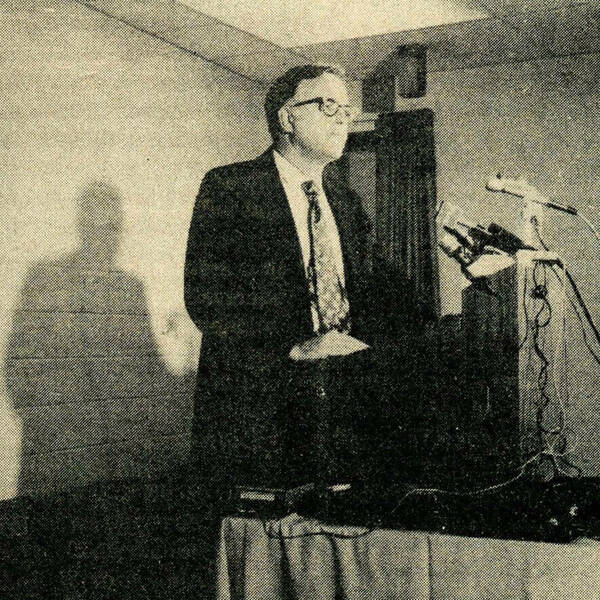 A man in a dark suit, light shirt, and patterned tie, wearing glasses, stands speaking at a podium with multiple microphones. He appears serious, with his hands clasped.